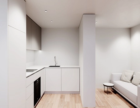 Helle, moderne und barrierefreie Wohnung. Links ist eine weiße Küche zu sehen und rechts ein kleiner Wohnbereich mit grauem Sofa.