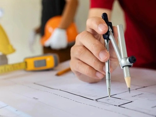 Ein Bauherr sitzt an einem Tisch und zeichnet mit einem Zirkel etwas in einen Bauplan.